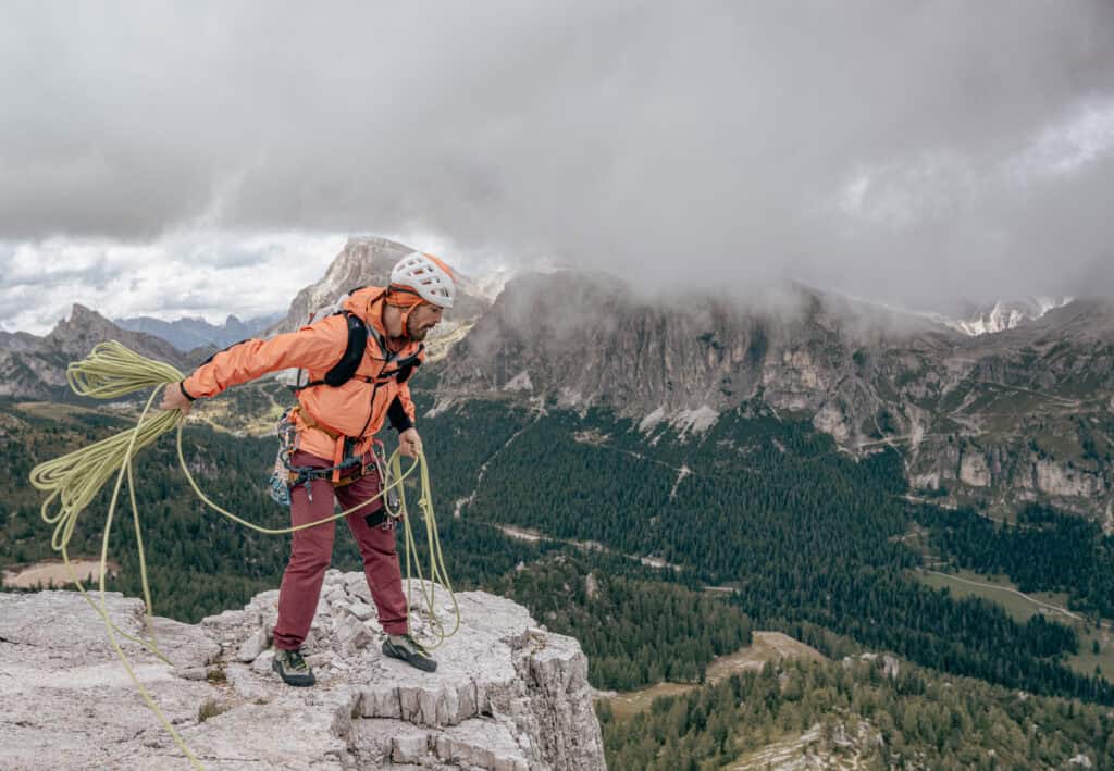 A climber is getting ready to abseil. He is throwing half-ropes down the rock face. Mountain scenery in the background